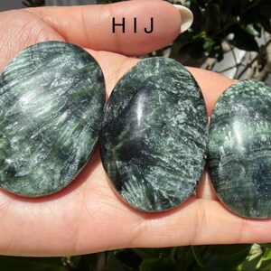 May include: Three oval-shaped green stones with dark green and black veining are held in a hand. The stones have a polished, smooth surface. The stones are a natural product.