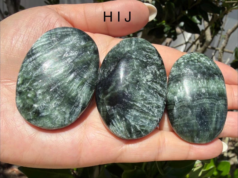 May include: Three oval-shaped green stones with dark green and black veining are held in a hand. The stones have a polished, smooth surface. The stones are a natural product.