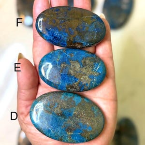 May include: Three polished, oval-shaped stones with a mix of blue and gold colors are held in a hand. The stones have a smooth, glossy surface. Additional stones are visible in the background. A blue beaded bracelet is visible on the wrist.