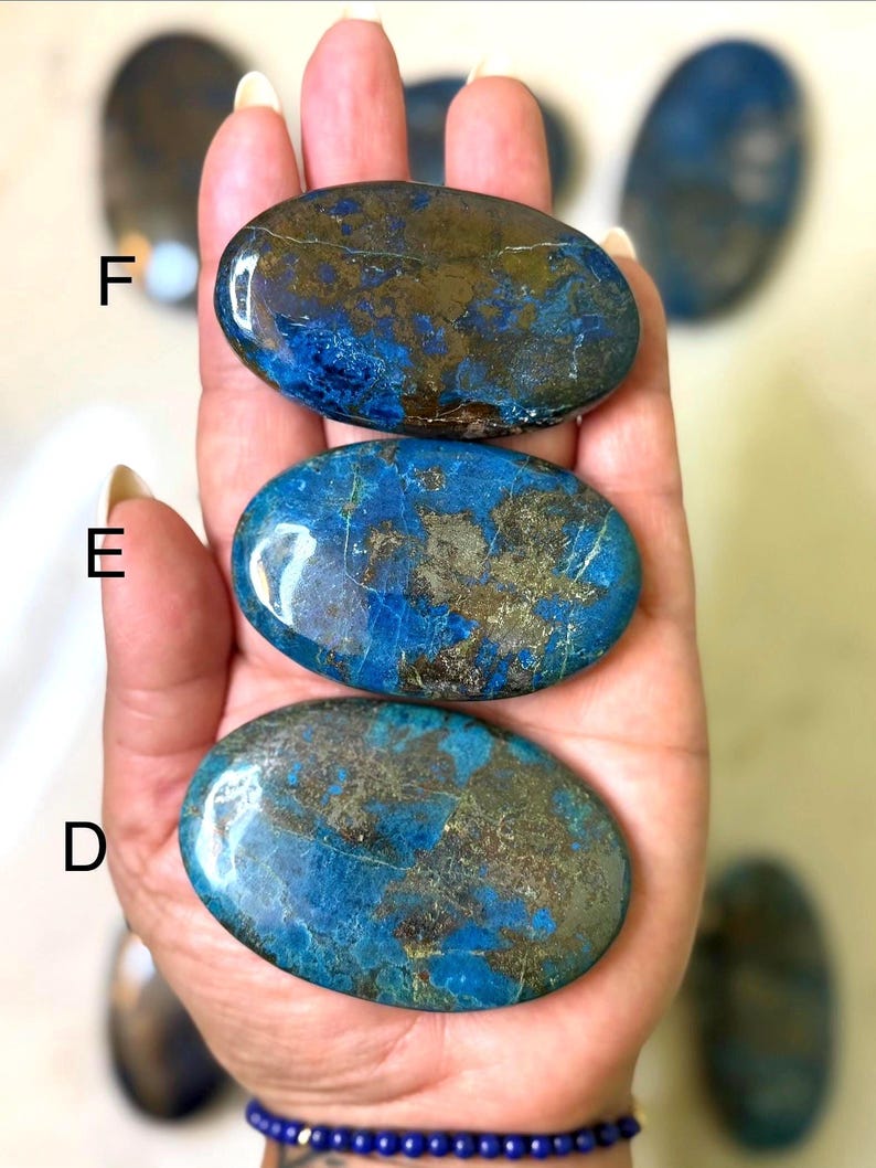May include: Three polished, oval-shaped stones with a mix of blue and gold colors are held in a hand. The stones have a smooth, glossy surface. Additional stones are visible in the background. A blue beaded bracelet is visible on the wrist.
