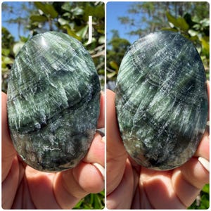May include: Two polished, oval-shaped stones with a swirling pattern of dark green and white. The stones are held in a person's hands, with the background showing green foliage and sunlight.