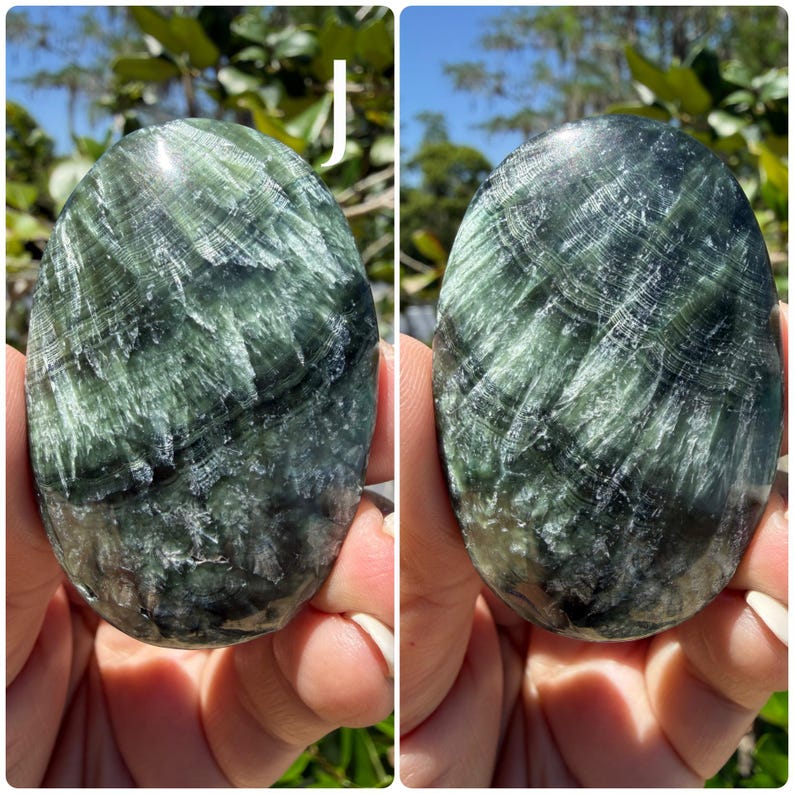 May include: Two polished, oval-shaped stones with a swirling pattern of dark green and white. The stones are held in a person's hands, with the background showing green foliage and sunlight.