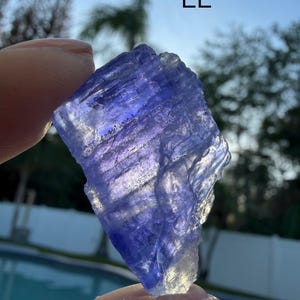 May include: A raw, translucent tanzanite crystal with a vibrant violet-blue hue and natural jagged edges. The stone displays internal striations and crystalline textures, catching the light to reveal deep purple and blue tones. Marked with the text EE.