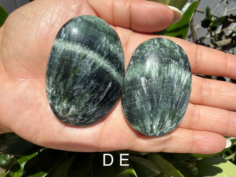 May include: Two oval-shaped, polished stones with a dark green and white striped pattern. The stones are smooth and appear to be natural gemstones. The image is well-lit, showcasing the details of the stones.