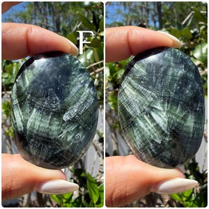 May include: Two oval-shaped, polished stones. The stones have a dark green base color with lighter green, fibrous patterns throughout. The stones are held between fingers against a blurred green background.