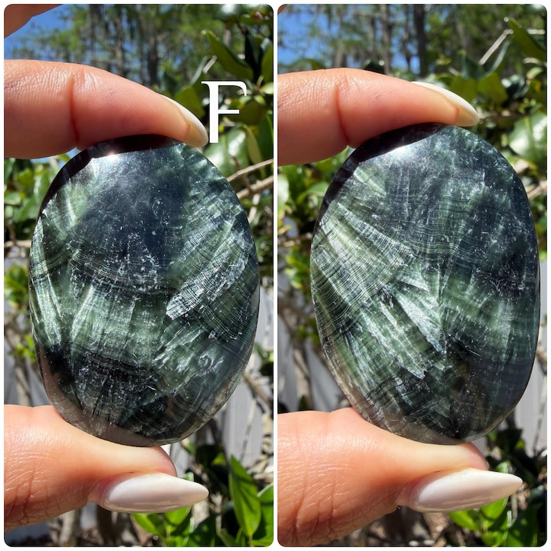 May include: Two oval-shaped, polished stones. The stones have a dark green base color with lighter green, fibrous patterns throughout. The stones are held between fingers against a blurred green background.