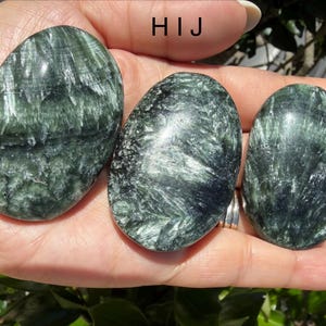 May include: Three oval-shaped seraphinite stones with a dark green color and a feathery, silvery pattern. The stones are smooth and polished, with a natural, organic appearance. The text "HIJ" is visible in the image.
