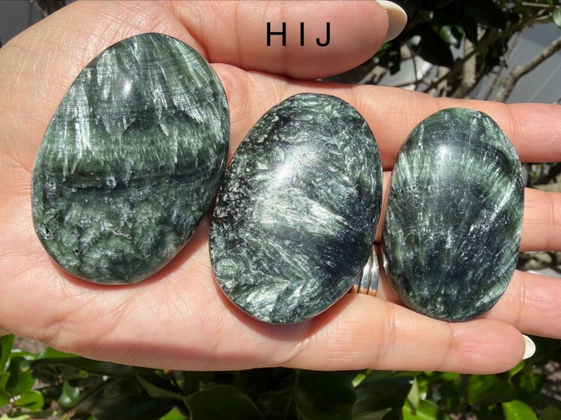 May include: Three oval-shaped seraphinite stones with a dark green color and a feathery, silvery pattern. The stones are smooth and polished, with a natural, organic appearance. The text "HIJ" is visible in the image.