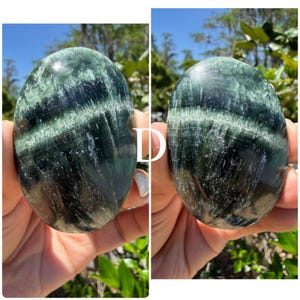 May include: Two polished, oval-shaped stones. The stones have a dark green base color with lighter green and white veining. The stones are held up against a background of green foliage and a blue sky.