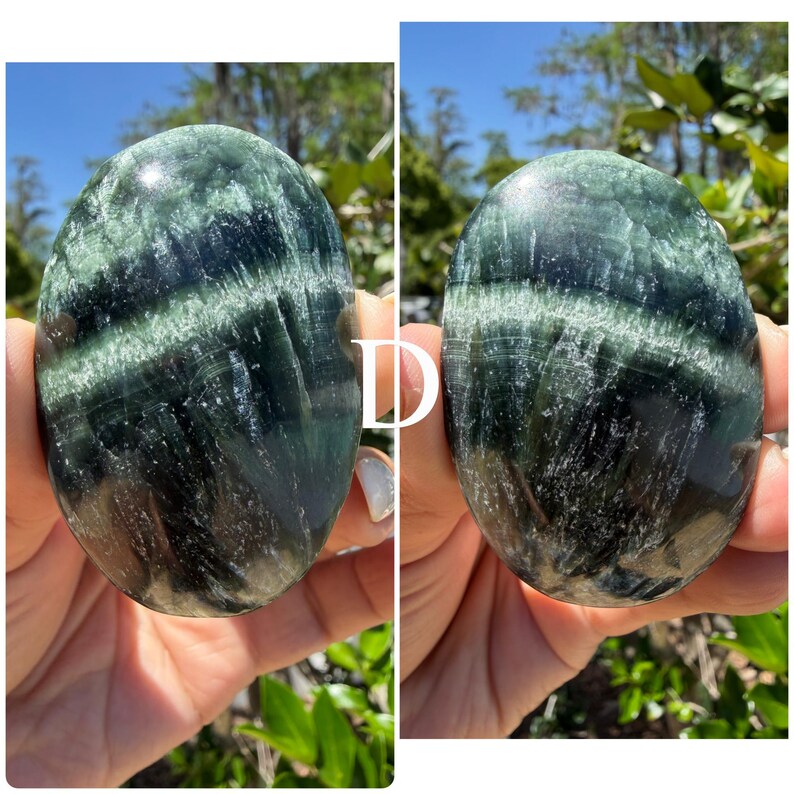 May include: Two polished, oval-shaped stones. The stones have a dark green base color with lighter green and white veining. The stones are held up against a background of green foliage and a blue sky.
