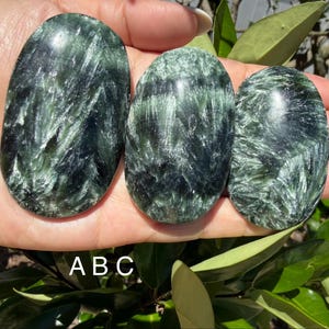 May include: Three polished seraphinite stones with a dark green and silver feathery pattern. The stones are oval-shaped and held in a hand, with the letters "ABC" visible below. The stones are displayed against a backdrop of green foliage.