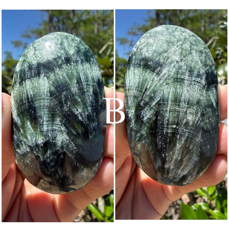 May include: Two polished oval-shaped stones. The stones have a swirling pattern of dark green and black, with a glossy finish. The stones are held in a person's hands, with a blurred background of greenery and blue sky.