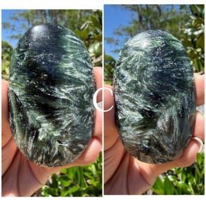 May include: Two polished seraphinite stones, each held in a hand, are displayed side-by-side. The stones are oval-shaped with a dark green base color and feathery, silvery-white inclusions. The stones are smooth and reflect light.