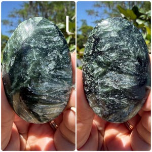 May include: Two polished serpentine stones, each with a dark green and white marbled pattern. The stones have an oval shape and a smooth, glossy surface. The background is blurred, with hints of green foliage and blue sky.