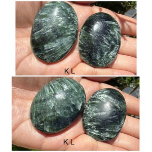 May include: Four oval-shaped, polished green stones with a swirling pattern are displayed. The stones are held in a person's hand. The text "KL" is visible on the image. These stones are likely used for healing or decorative purposes.