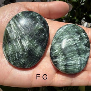 May include: Two polished, oval-shaped green stones with a swirling, textured pattern. The stones are held in a hand, showcasing their smooth, reflective surfaces. The letter "F G" is visible at the bottom of the image.