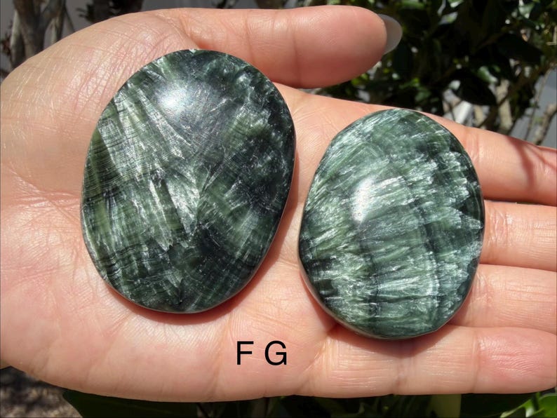 May include: Two polished, oval-shaped green stones with a swirling, textured pattern. The stones are held in a hand, showcasing their smooth, reflective surfaces. The letter "F G" is visible at the bottom of the image.