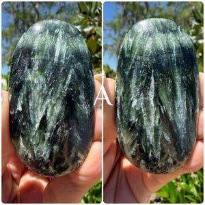 May include: Two oval-shaped Seraphinite stones. The stones have a dark green base color with feathery, silvery-white inclusions. The polished stones are held up against a background of green foliage and a bright blue sky.