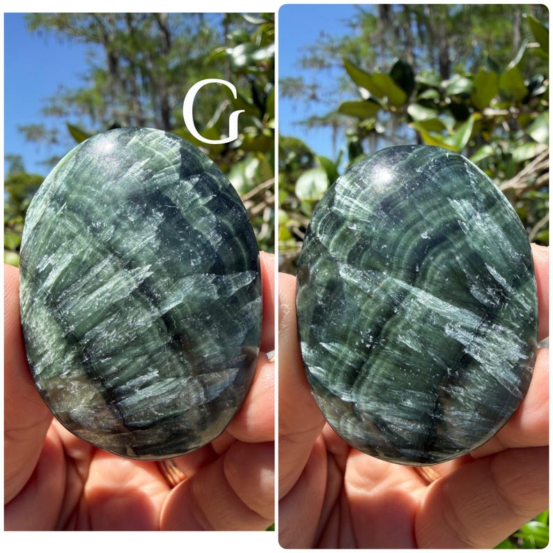 May include: Two oval-shaped green Serpentine stones with a swirling pattern of dark green and white veins. The stones are held in a person's hands, with a blurred background of trees and sky.