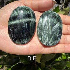 May include: Two oval-shaped green Serpentine stones with dark green and white veining. The stones are smooth and polished, with a glossy finish. The stones are held in a hand, with the letter "DE" visible at the bottom.
