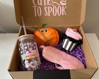 Puede incluir: Una caja de regalo con temática de Halloween con un vaso transparente, un artículo con forma de calabaza, una máscara para los ojos y un artículo con forma de cupcake. La caja tiene el texto "TOO CUTE TO SPOOK".