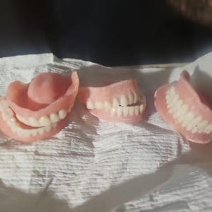 Puede incluir: Tres juegos de dentaduras postizas de acr&iacute;lico rosa con dientes blancos dispuestos en fila. El juego de la izquierda muestra los arcos superior e inferior, el del medio muestra un arco parcial y el de la derecha muestra un solo arco.