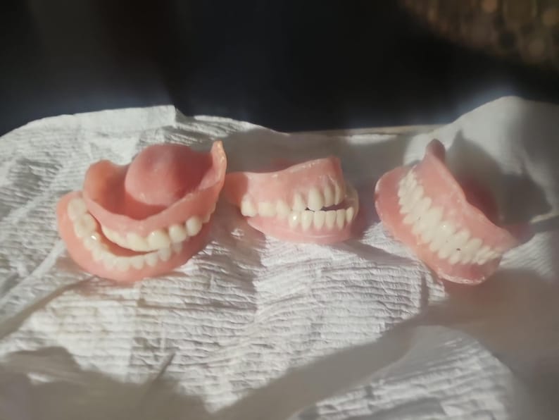 Puede incluir: Tres juegos de dentaduras postizas de acr&iacute;lico rosa con dientes blancos dispuestos en fila. El juego de la izquierda muestra los arcos superior e inferior, el del medio muestra un arco parcial y el de la derecha muestra un solo arco.
