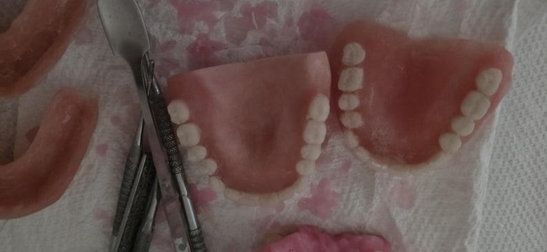Puede incluir: Dos juegos de dentaduras postizas de acr&iacute;lico rosa con dientes blancos dispuestos sobre una superficie texturizada junto a herramientas dentales de metal. Las dentaduras presentan una base curva color enc&iacute;a con dientes blancos.