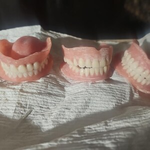 Puede incluir: Tres juegos de dentaduras postizas con bases de enc&iacute;a de acr&iacute;lico rosa y dientes artificiales blancos. Las dentaduras est&aacute;n dispuestas en fila, mostrando varias configuraciones de arcos dentales con formas de dientes y enc&iacute;as realistas.