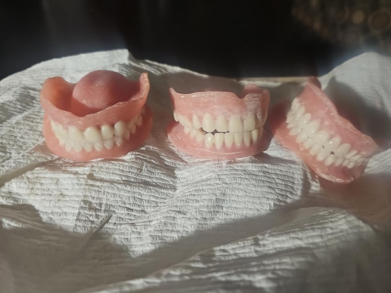 Puede incluir: Tres juegos de dentaduras postizas con bases de enc&iacute;a de acr&iacute;lico rosa y dientes artificiales blancos. Las dentaduras est&aacute;n dispuestas en fila, mostrando varias configuraciones de arcos dentales con formas de dientes y enc&iacute;as realistas.