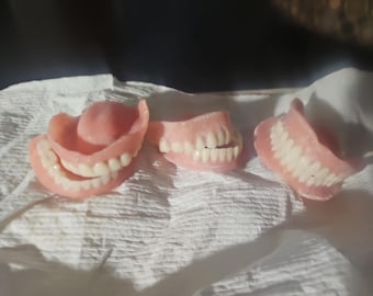 Puede incluir: Tres juegos de dentaduras postizas de acrílico rosa con dientes blancos dispuestos en fila. El juego de la izquierda muestra los arcos superior e inferior, el del medio muestra un arco parcial y el de la derecha muestra un solo arco.