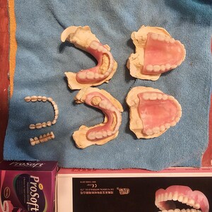 Puede incluir: Una colecci&oacute;n de pr&oacute;tesis dentales que incluye cuatro dentaduras parciales y completas fijadas en moldes de yeso, dos segmentos de puente dental separados y dos cajas de productos etiquetadas Prosoft y Cai Yu Dental Materials Co., Ltd. con un modelo.
