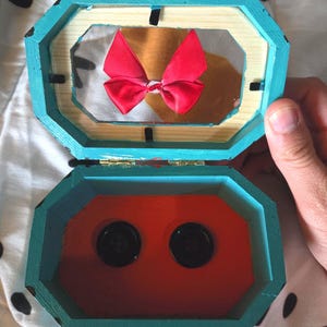 Puede incluir: Una caja de madera azul turquesa con un interior rojo. La caja est&aacute; abierta y tiene un lazo rojo en el interior. La caja tiene dos botones negros en la parte inferior.