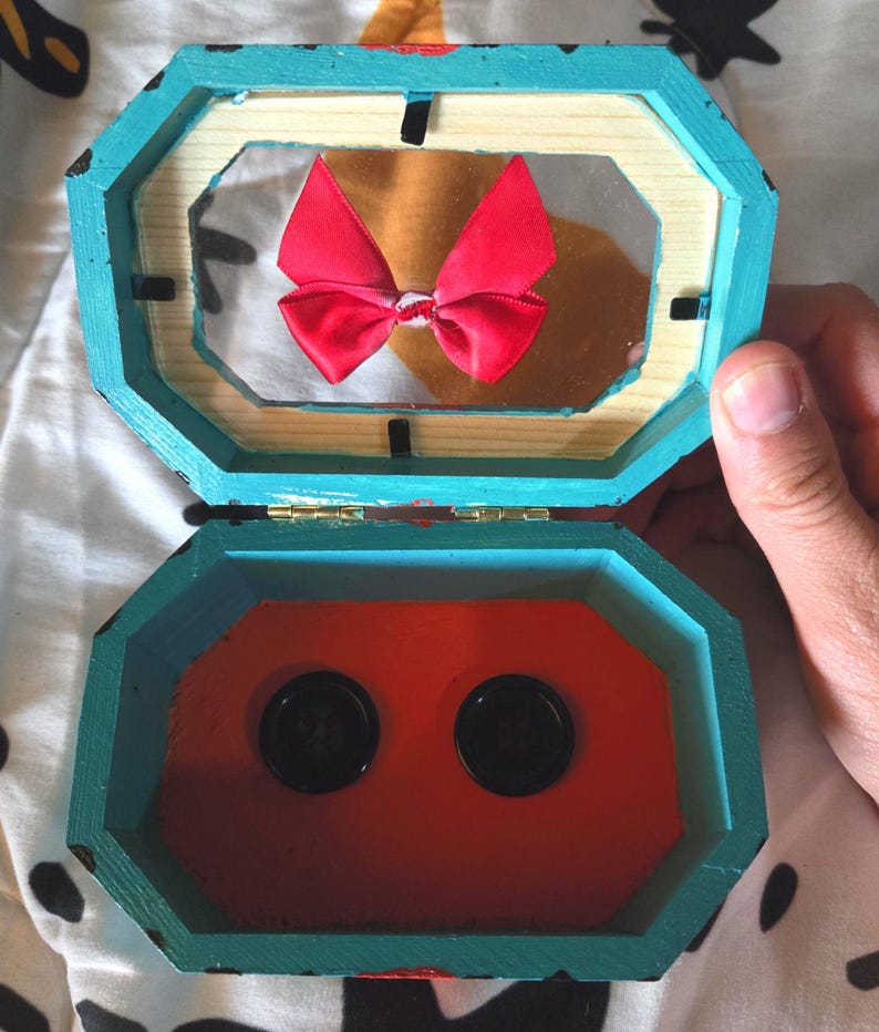 Puede incluir: Una caja de madera azul turquesa con un interior rojo. La caja est&aacute; abierta y tiene un lazo rojo en el interior. La caja tiene dos botones negros en la parte inferior.