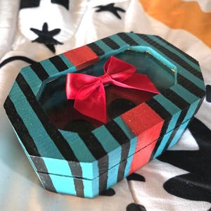 Puede incluir: Una peque&ntilde;a caja de madera octogonal con un patr&oacute;n de rayas turquesa, negro y rojo. La caja tiene una ventana de vidrio transparente en la parte superior, que revela un lazo rojo en el interior.