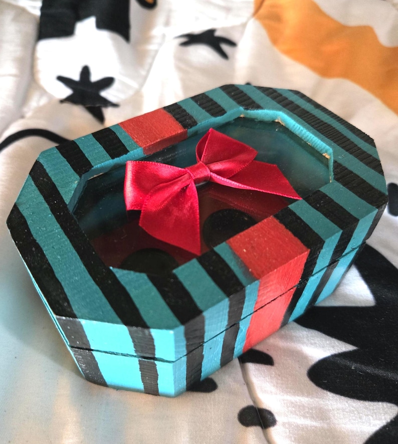 Puede incluir: Una peque&ntilde;a caja de madera octogonal con un patr&oacute;n de rayas turquesa, negro y rojo. La caja tiene una ventana de vidrio transparente en la parte superior, que revela un lazo rojo en el interior.