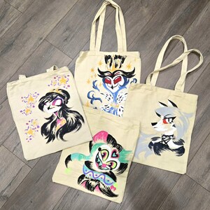 May include: Four cream-colored canvas tote bags, each featuring a unique hand-painted character illustration. The designs include a dark-haired figure with stars, a blue-feathered owl-like character, a colorful jester-like figure, and a grey wolf-like character.