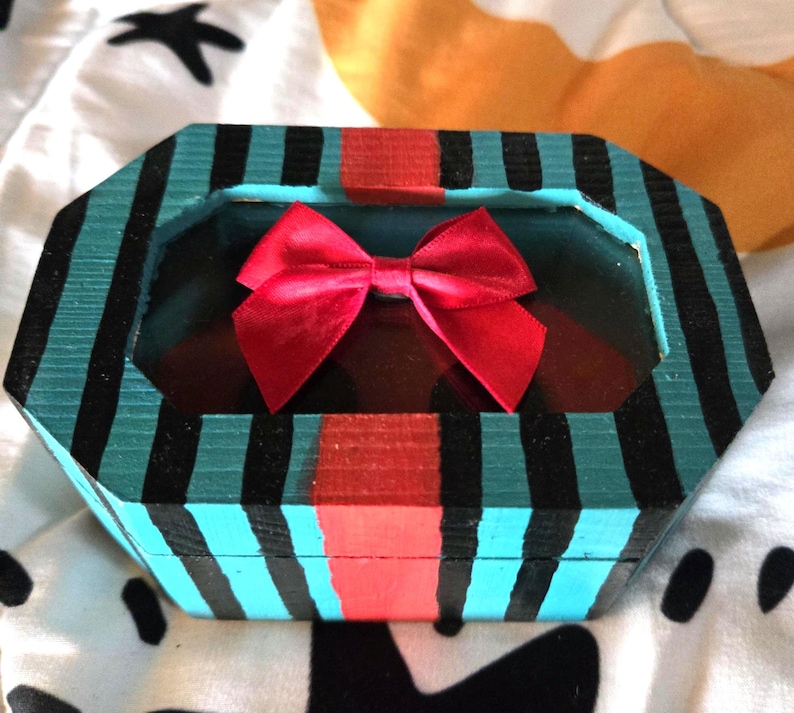 Puede incluir: Una caja de madera a rayas azul turquesa y negras con una cinta roja y una ventana de vidrio transparente. La caja est&aacute; decorada con una franja roja en el centro.