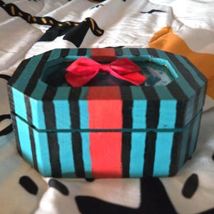 Puede incluir: Una peque&ntilde;a caja de madera octogonal con un dise&ntilde;o a rayas azul verdoso y negro. La caja tiene una cinta roja y una tapa de vidrio transparente.