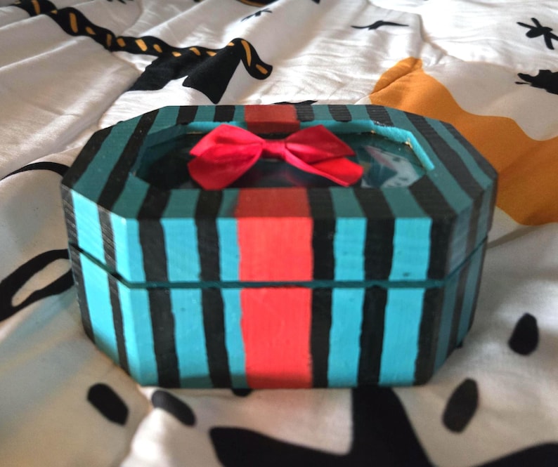 Puede incluir: Una peque&ntilde;a caja de madera octogonal con un dise&ntilde;o a rayas azul verdoso y negro. La caja tiene una cinta roja y una tapa de vidrio transparente.