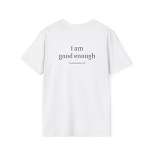 以下が含まれることがあります： 背面に「I am good enough」と「ItsAWoofWorld」の文字がプリントされた白いTシャツです。