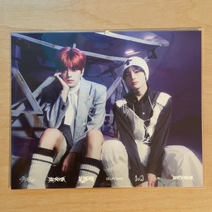 May include: Two men wearing casual clothing pose for a photo. The man on the left is wearing a gray plaid blazer, white shirt, and black and white striped socks. The man on the right is wearing a white beanie, a white shirt with a black lace overlay, and white pants. The photo is signed by the men, "Seungmin" and "I.N."  The photo is also signed with the band name "Stray Kids".