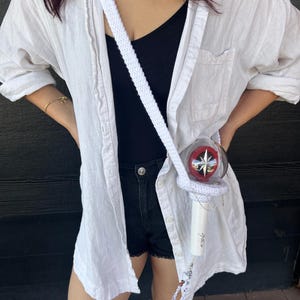 May include: A white crochet crossbody bag with a strap, holding a clear and red light stick. The light stick has a star design and the text "Stray Kids". The words "Crochet Pattern" are at the bottom of the image.