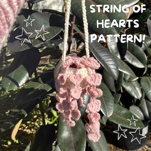 May include: A pink crocheted string of hearts plant hanger with a white crocheted pot. The hanger is hanging from a white cord and is in front of a green leafy plant. The text "STRING OF HEARTS PATTERN!" is written in white on a black background above the hanger.