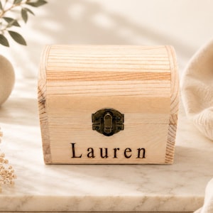 May include: A light-colored wooden chest with a curved lid and a dark metal latch. The name Lauren is engraved in a dark serif font on the front panel of the box. The wood grain is visible across the surface of the container.