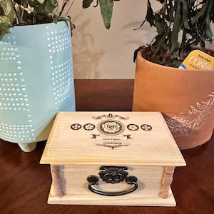 May include: A wooden box with a hinged lid and a metal handle. The box is engraved with the words "Roe Lynn Collection" and a floral design.