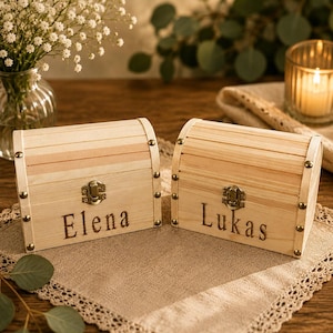 May include: Two light-colored wooden chest-style boxes with arched lids and metal latches. Each box features a decorative metal stud at each corner and is engraved with a name on the front, one reading Elena and the other reading Lukas.