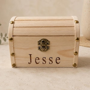 May include: A light-colored wooden chest with a curved lid, featuring a metal latch in the center and decorative brass-toned studs on the corners. The name Jesse is engraved in a dark serif font on the front panel of the wooden box.
