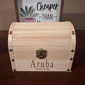 Puede incluir: Un pequeño cofre de madera clara con la parte superior curvada y detalles metálicos. La palabra "Aruba" y el año "2025" están grabados en la parte delantera. Un letrero enmarcado en el fondo dice "Cheaper -THAN-" con una ilustración de planta.