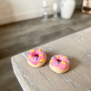 May include: Two miniature donuts with pink icing and colourful sprinkles. The donuts have a light brown base and are placed on a light-coloured wooden surface. The sprinkles are yellow and blue.